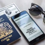 Flat lay with UK passport, American Express card, smartphone showing insurance policy, and sunglasses on grey marble background