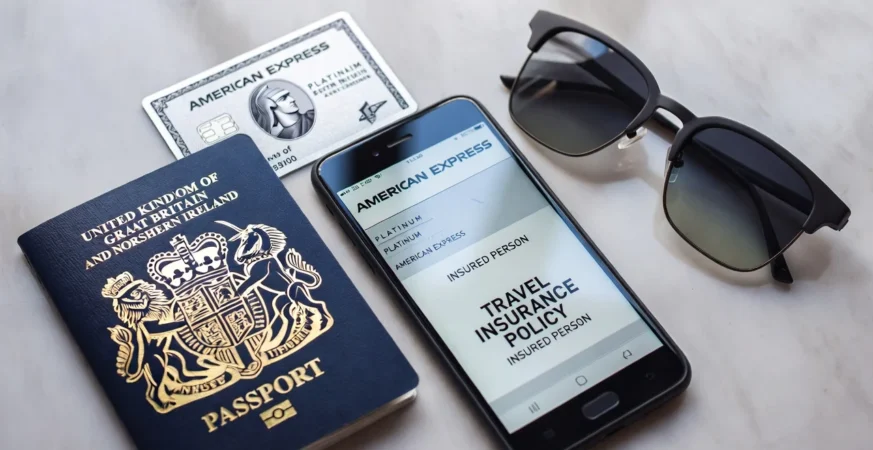Flat lay with UK passport, American Express card, smartphone showing insurance policy, and sunglasses on grey marble background