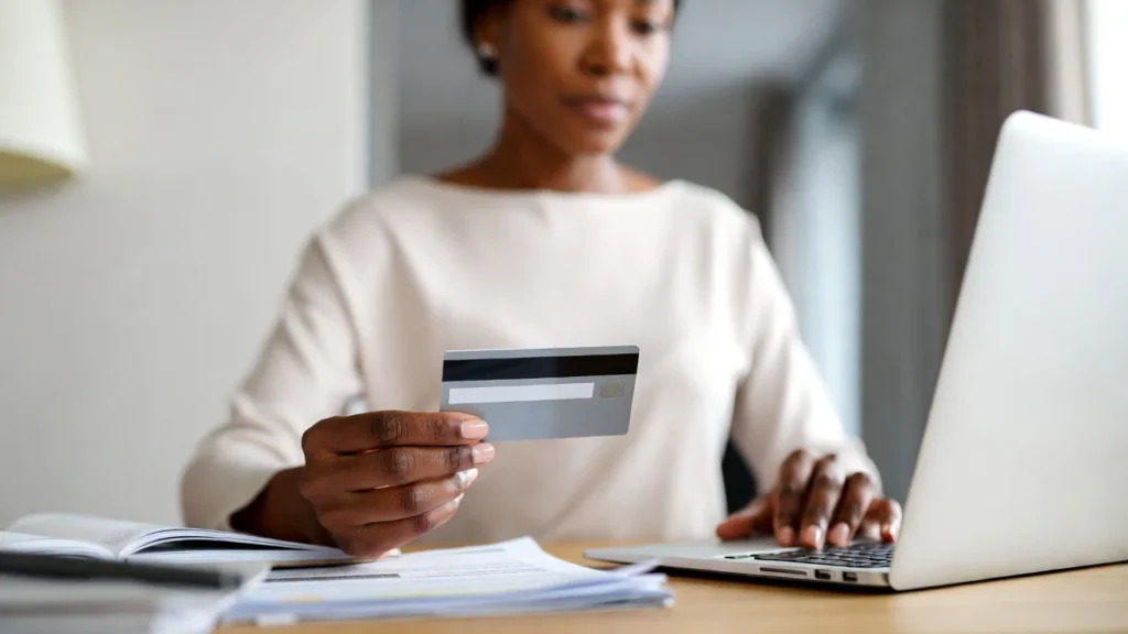 Person holding credit card while reviewing financial documents on laptop in UK home setting