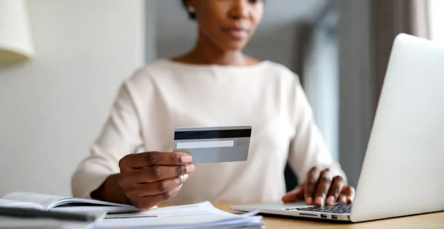 Person holding credit card while reviewing financial documents on laptop in UK home setting